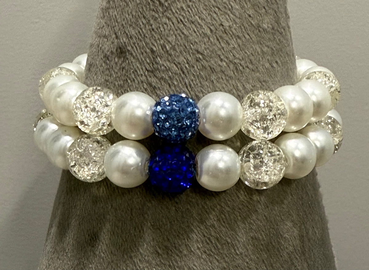 A pair of beaded stretch bracelets displayed on a gray cone stand. The bracelets feature white faux pearls, clear faceted crystal beads, and two prominent deep blue glitter beads.