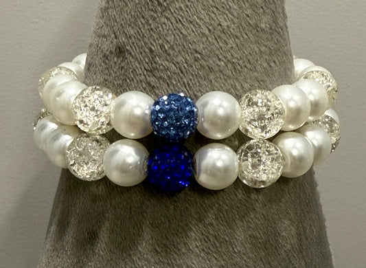 A pair of beaded stretch bracelets displayed on a gray cone stand. The bracelets feature white faux pearls, clear faceted crystal beads, and two prominent deep blue glitter beads.