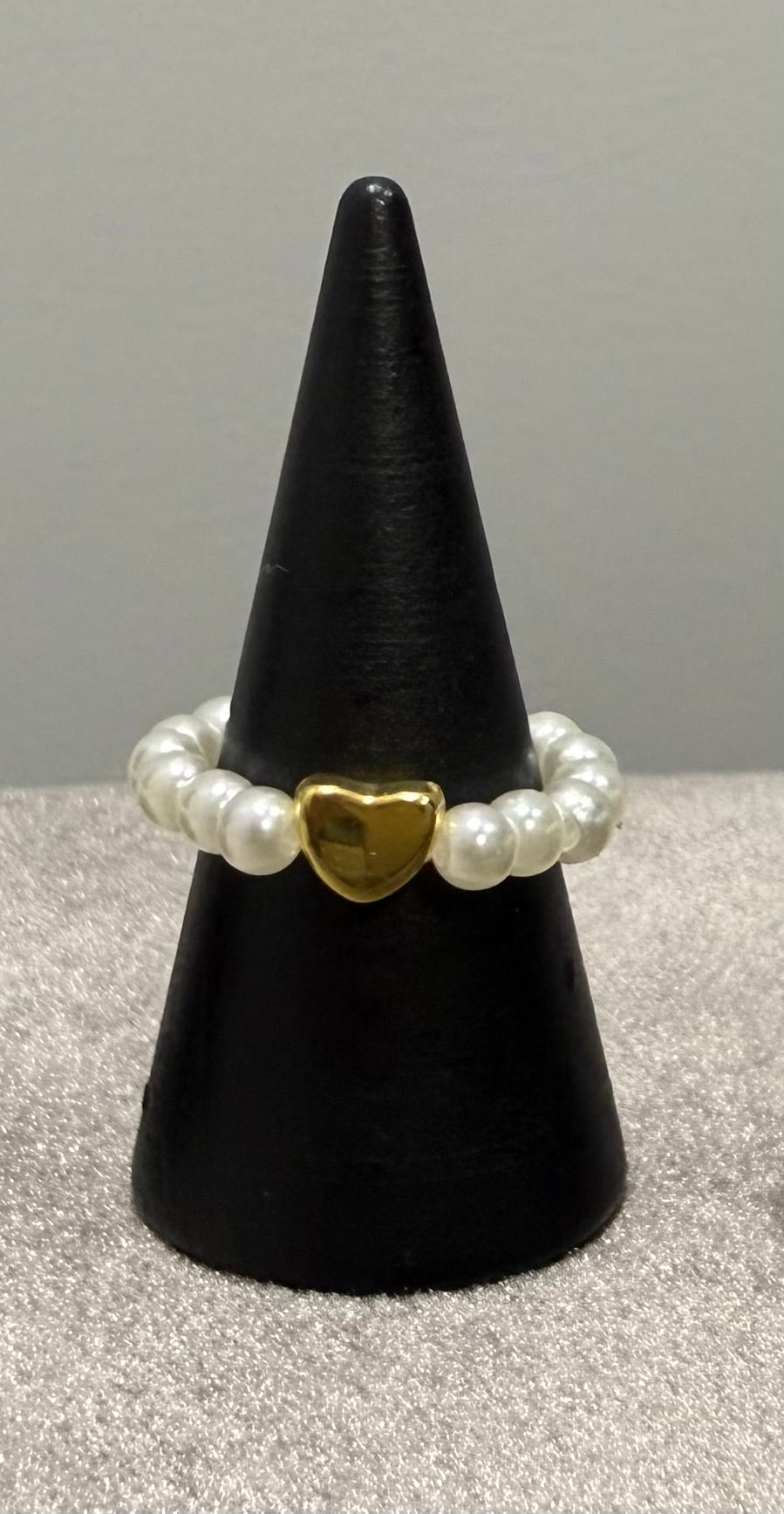 A beaded stretch bracelet with white pearl-like beads and a central gold heart charm, displayed on a black cone stand against a neutral gray background.