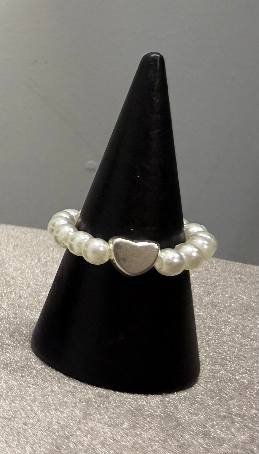 A beaded stretch ring with white pearl-like beads and a central silver heart charm, displayed on a black cone stand against a gray background.