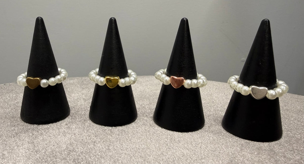 Four pearl beaded rings with heart-shaped metal accents displayed on black cone stands. The heart accents are gold, yellow gold, rose gold, and silver, set against a neutral gray background.