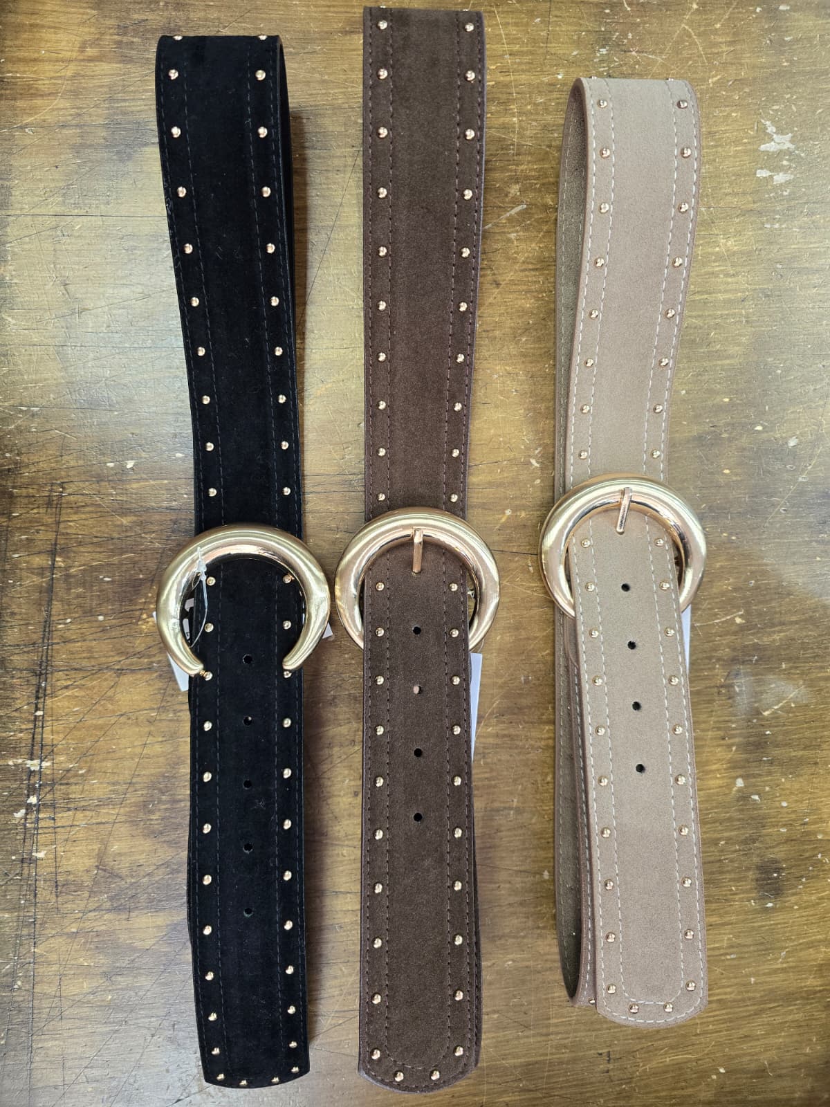 Three suede leather belts in black, brown, and gray with silver rings on a wooden surface.