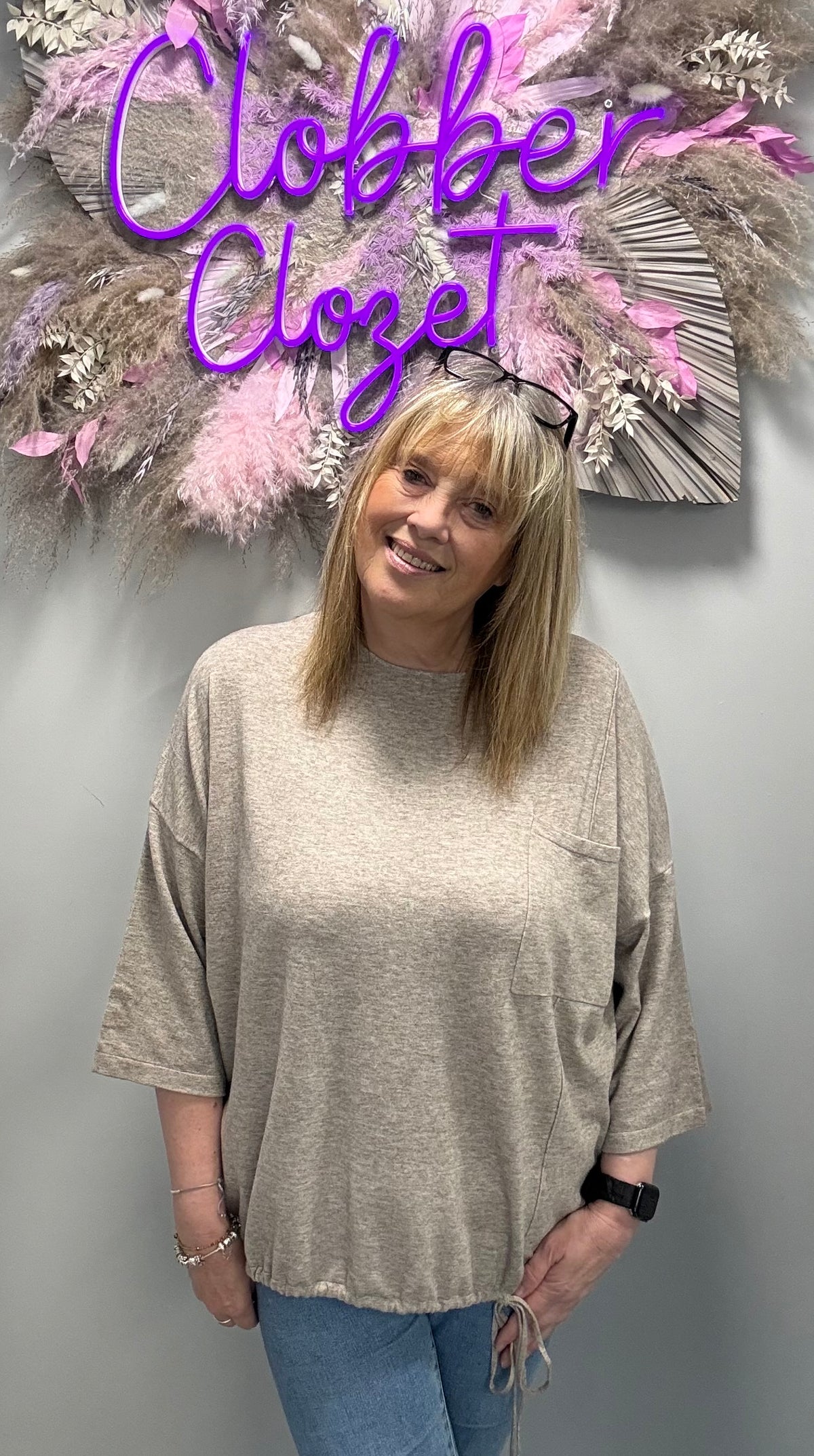 A woman wearing a loose, oversized short-sleeve knit top in a heathered light taupe-beige color, standing in front of a gray wall with a decorative floral wall hanging and the text 'Clobber Closet' in purple script.