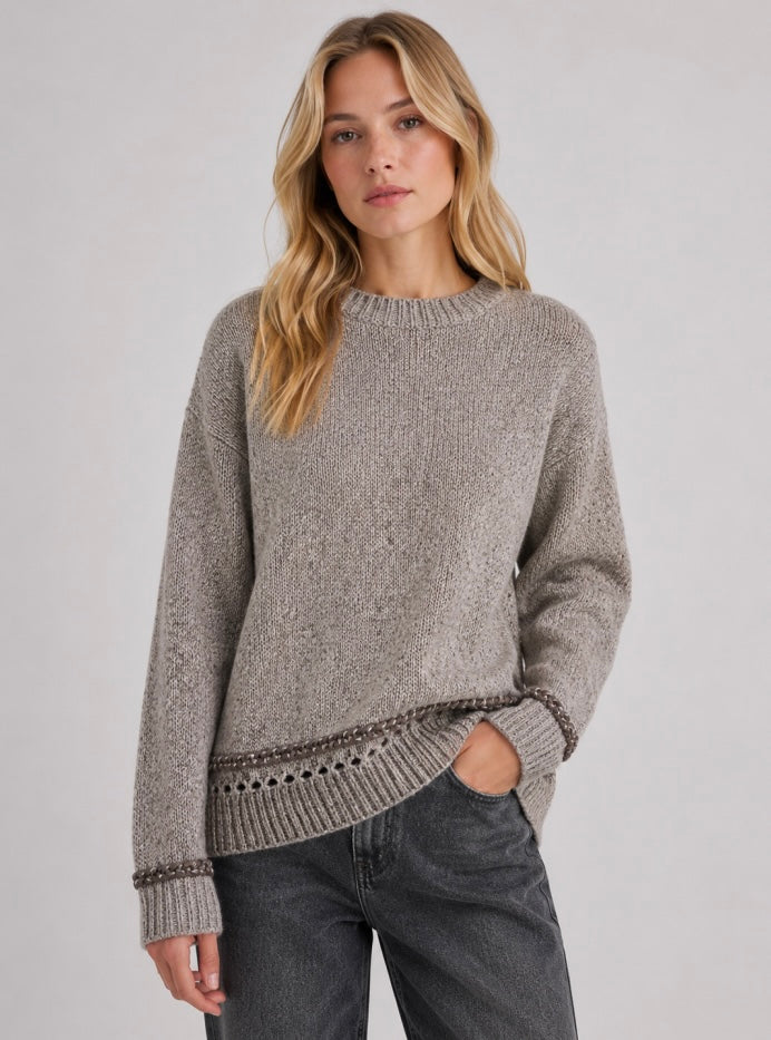 A woman wearing a taupe knit sweater with a textured pattern and decorative ribbed hem, paired with dark gray jeans, against a plain light gray studio background.