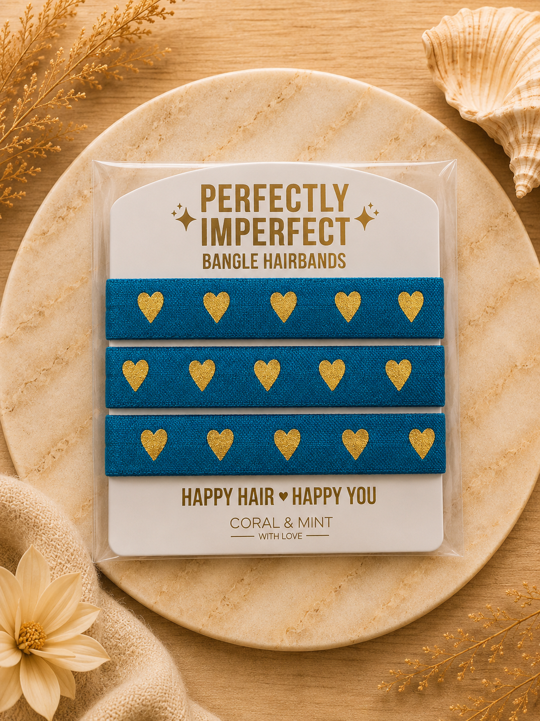 Three blue bangle hairbands with gold heart patterns, packaged on a white card that reads 'PERFECTLY IMPERFECT BANGLE HAIRBANDS' and 'HAPPY HAIR HAPPY YOU', displayed on a round wooden tray with decorative dried flowers and a seashell.