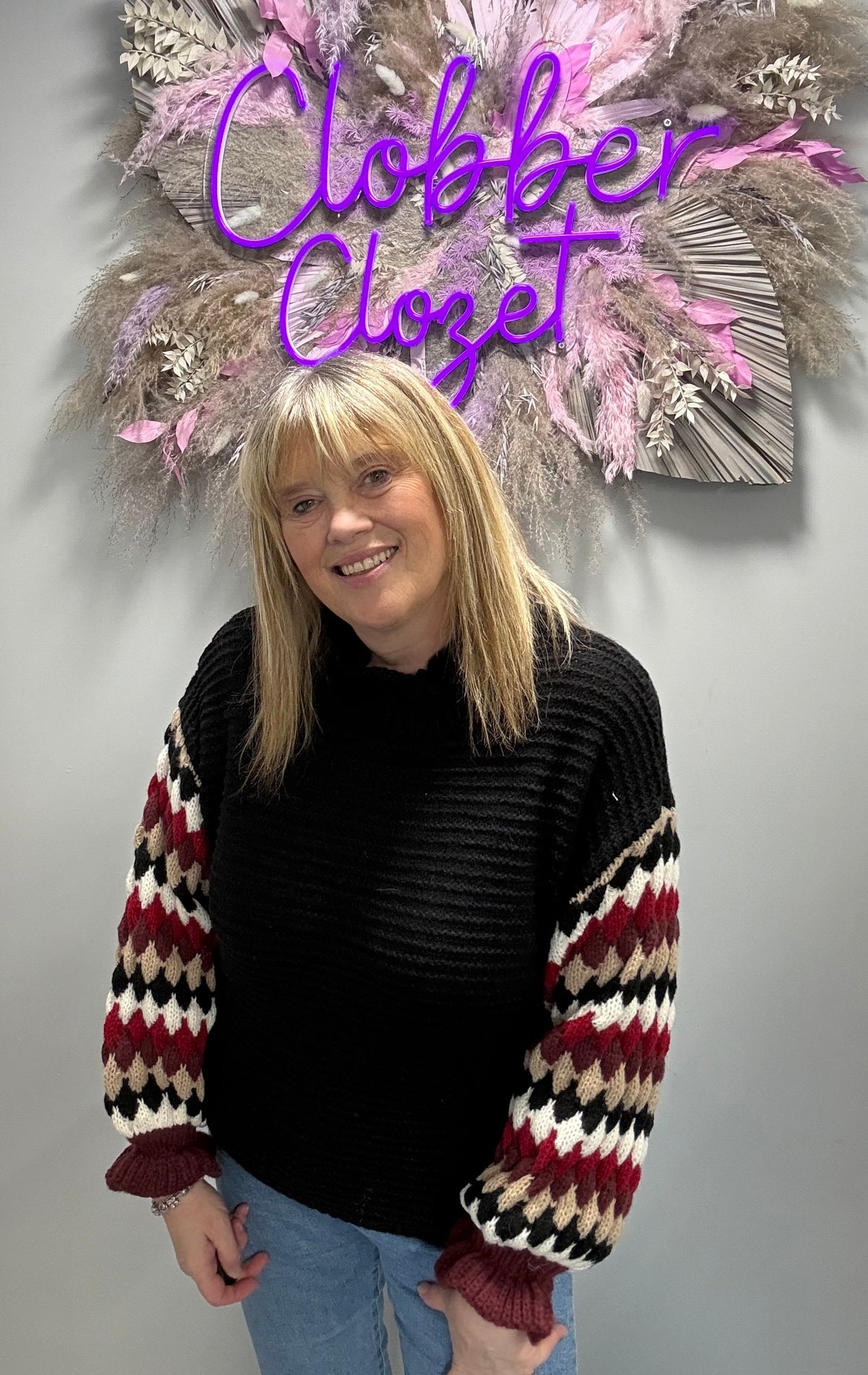 A woman with blonde hair wearing a black ribbed knit sweater with patterned sleeves, standing in front of a gray wall with a decorative dried floral arrangement and a purple neon sign that reads 'Clobber Closet'.