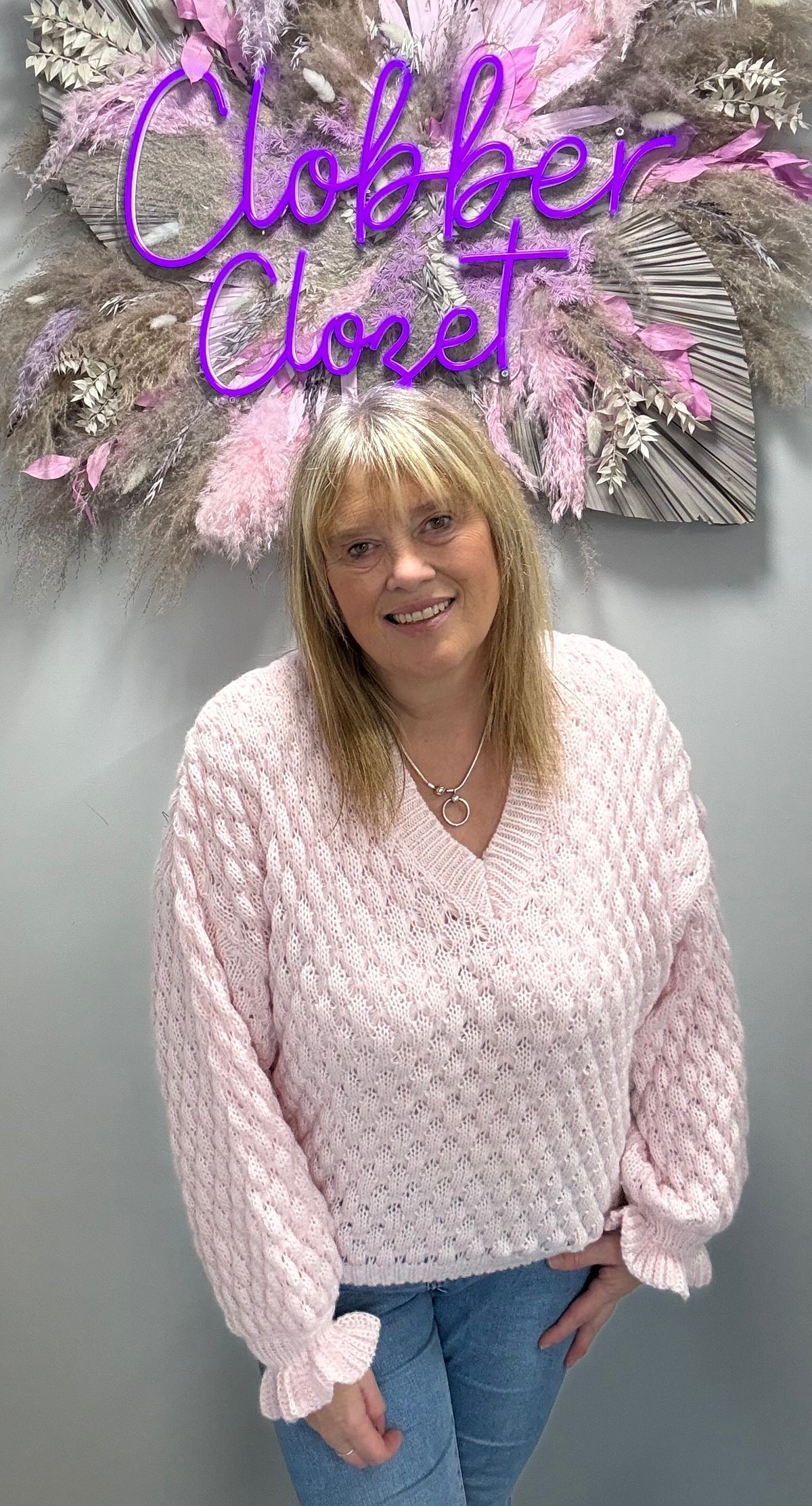 A woman wearing a light pink chunky cable-knit sweater with ruffled cuffs, standing in front of a gray wall with a decorative floral arrangement and a purple 'Closet' sign.