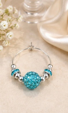 A silver-tone wine glass charm with a large, round, sparkling turquoise blue bead at the center, flanked by two smaller matching blue beads and silver spacer beads, displayed on a beige fabric background.