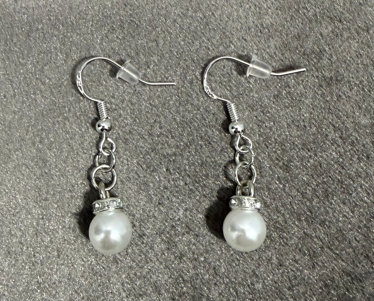 A pair of silver drop earrings with white pearl-like beads and small clear rhinestone accents, displayed on a textured gray background.