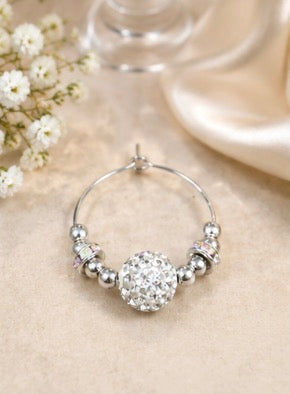 A silver wine glass charmwith a large, round, crystal-encrusted center and small silver beads along the side, displayed on a beige fabric surface with soft decorative props.