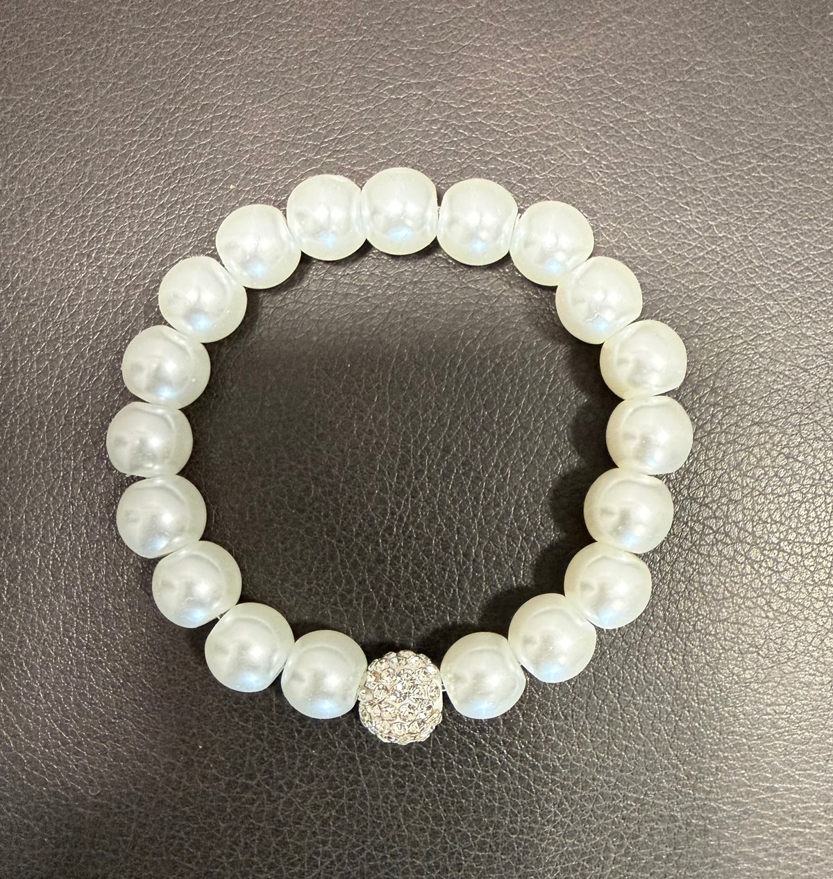 A beaded stretch bracelet featuring round white faux pearls and a textured silver-toned round charm, displayed on a dark gray textured leather surface.