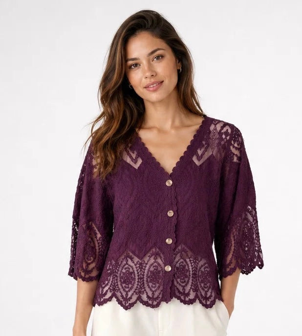 A woman wearing a dark purple lace cardigan with scalloped edges and three-quarter sleeves, paired with white pants, against a plain white studio background.