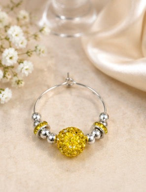 A silver wind glass charm with a large textured yellow center bead and smaller yellow and silver accent beads, displayed on a beige fabric surface with small white flowers and a satin ribbon in the background.