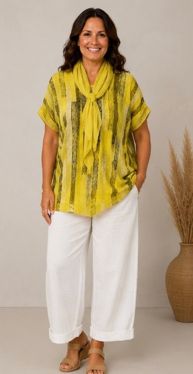 A woman modeling a yellow and olive green patterned short-sleeve top with a matching tie-neck scarf, paired with white wide-leg pants and tan sandals, standing in front of a neutral beige background.